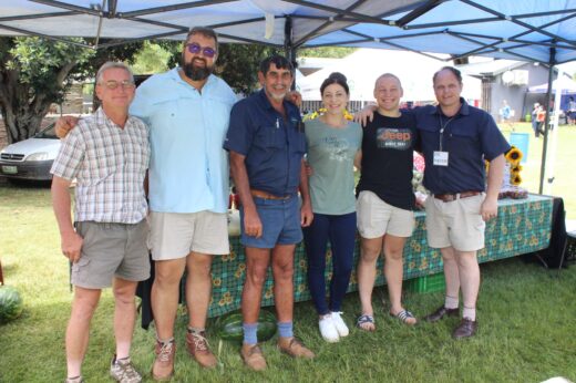 A group of six people stands smiling under a blue canopy at an outdoor event. They are casually dressed and standing on grass with a table behind them.
