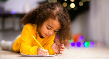 A young child with curly hair in a yellow sweater lies on a carpet, focused on drawing with a pencil. Soft, colorful lights create a warm, festive atmosphere.