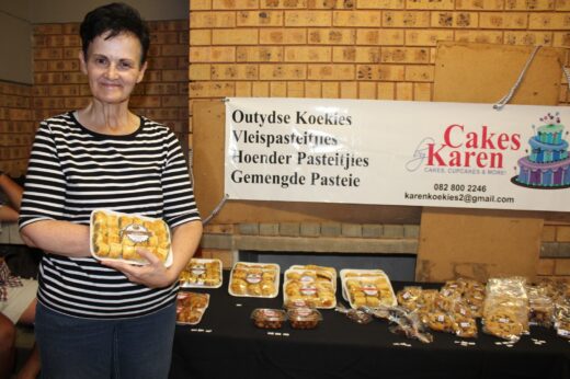 A woman in a striped shirt holds a tray of baked goods in front of a table displaying various pastries. A banner reads "Cakes by Karen" on the wall behind her.