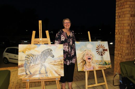 A woman stands smiling between two paintings on easels. One depicts a zebra in a savanna, and the other shows a laughing child with a windmill. Evening setting.