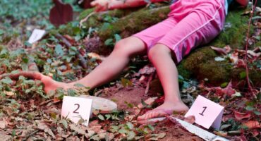 The image depicts a crime scene in a forest. A person lies on the ground wearing pink shorts, surrounded by evidence markers and a knife. The tone is tense and mysterious.