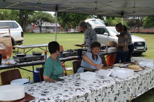 A group of people under a tent preparing food at an outdoor event. They look cheerful and engaged, with hot plates and food containers on the tables.