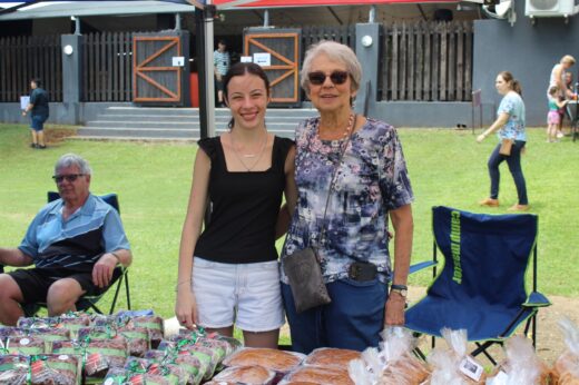 A young woman and older woman smile at a market stall displaying packaged bread. An older man sits nearby in a chair on a sunny day, conveying a cheerful atmosphere.