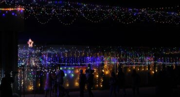 Outdoor scene at night with colorful string lights hanging above, creating a festive atmosphere. Silhouettes of people admire the display.