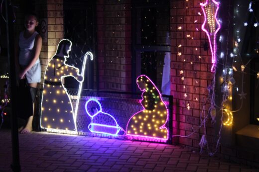 Illuminated nativity scene with vibrant lights depicts Mary, Joseph, and baby Jesus on a porch. A child stands nearby, creating a warm, festive atmosphere.