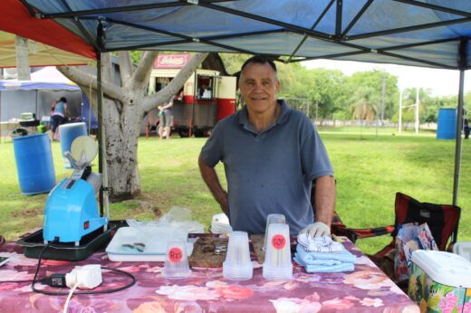 A friendly man stands under a canopy at an outdoor market stall. A shaved ice machine and stacked cups are on a floral tablecloth. The background is grassy with trees and tents, conveying a casual, lively atmosphere.