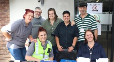 A group of seven people smiling at the camera, some are standing and others sitting at a table with cash boxes. A "No under 18" sign is in the background.