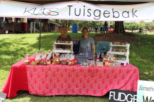 Two women stand behind a market stall labeled "Klits Tuisgebak," displaying homemade baked goods. The scene is outdoors, and the atmosphere is cheerful.
