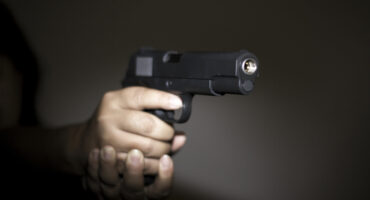 A hand firmly grips a black handgun in a dimly lit setting. The focus is on the barrel, creating a tense and serious atmosphere.