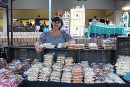 A woman with reindeer antlers stands behind a market stall displaying various packaged snacks. The scene is festive and busy, with shoppers in the background.