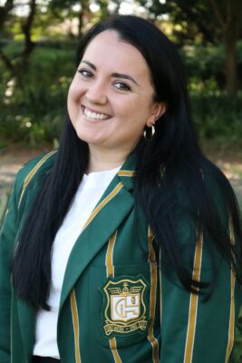 Smiling woman with long dark hair wearing a green and yellow striped blazer with an emblem. She stands outdoors with blurred trees and foliage.
