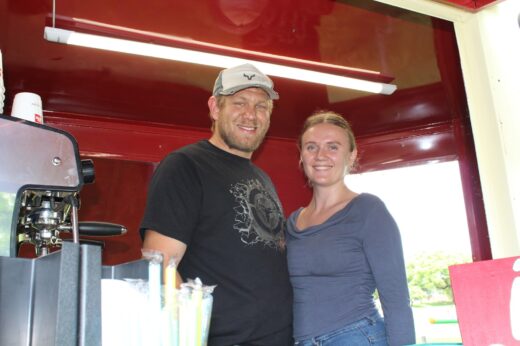 A smiling man and woman stand inside a red food truck with a coffee machine and colorful straws on the counter. Bright, welcoming atmosphere.