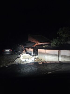 Dark night scene illuminated by headlights. A car is parked beside a fence, leading to a brick house. A tree fallen over a wall is visible.