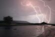 A dramatic sky illuminates with bright, jagged lightning over a dam. The landscape includes silhouetted hills and a lone tree, conveying a sense of awe and intensity.