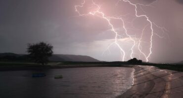 A dramatic sky illuminates with bright, jagged lightning over a dam. The landscape includes silhouetted hills and a lone tree, conveying a sense of awe and intensity.