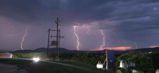 Tractor in a field under a dramatic sky with multiple vivid lightning strikes. Distant hills and an electric pole add depth. Intense and dynamic scene.