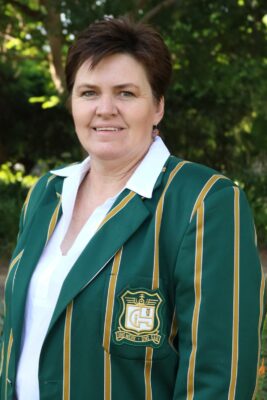 Woman in a green blazer with yellow stripes and a crest stands outside, smiling slightly. Background features lush greenery and soft sunlight.