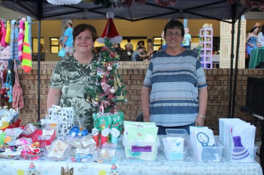 Two women smile behind a festively decorated market stall. The table displays handcrafted items, cards, and ornaments. A small Christmas tree adds a holiday touch.