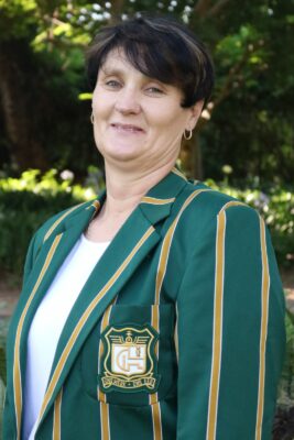 A woman in a green striped blazer with an emblem stands outdoors. She has short dark hair and smiles softly. Lush greenery frames the background.