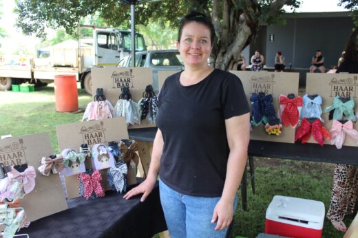 Smiling woman at outdoor market stall displaying colorful hair accessories with patterned bows. Background includes a tree, trucks, and seated people.