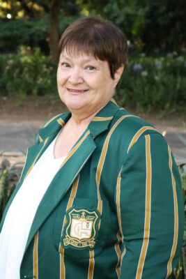 A woman with short brown hair smiles warmly, standing outdoors. She wears a green and gold striped blazer with a logo. The background is lush greenery.