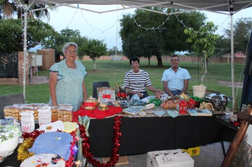A smiling older woman with two seated individuals at an outdoor market stall displaying homemade crafts and baked goods on a table. Festive atmosphere.