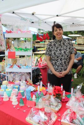 Smiling woman at a festive outdoor market booth with colorful handmade candles and gift baskets on display. White canopy tent, cheerful atmosphere.