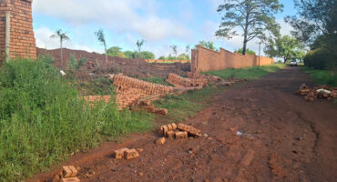 A red dirt road runs alongside a collapsed brick wall, with scattered bricks and green grass. Tall trees and a blue sky provide a calm backdrop.