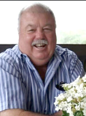 A cheerful man in a striped shirt laughs heartily, seated indoors. A vase of white flowers is in the foreground, adding a fresh, lively touch.