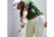 Golfer in mid-swing, wearing a green striped shirt, beige pants, and a white hat, focused and determined. Bright, sunny day backdrop.