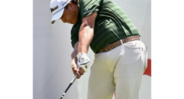 Golfer in mid-swing, wearing a green striped shirt, beige pants, and a white hat, focused and determined. Bright, sunny day backdrop.