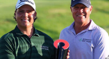 Two smiling men in golf attire, wearing caps, pose outdoors holding a trophy. They exude joy and achievement on a sunlit golf course background.