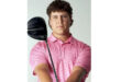A person holding a golf club, wearing a pink patterned shirt, stands confidently against a neutral background. The expression is focused and determined.