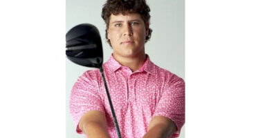 A person holding a golf club, wearing a pink patterned shirt, stands confidently against a neutral background. The expression is focused and determined.