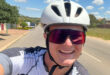 Smiling cyclist wearing a white helmet and sunglasses takes a selfie on a sunny day. Background shows a suburban street with greenery and houses.