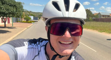 Smiling cyclist wearing a white helmet and sunglasses takes a selfie on a sunny day. Background shows a suburban street with greenery and houses.