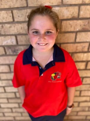 Smiling child with light brown hair in a ponytail, wearing a red polo shirt with a blue collar, stands against a brick wall. The mood is cheerful.