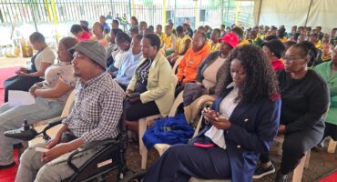 A diverse group of people is seated indoors, attentively listening to a presentation. The mood is focused and engaged. A man in a wheelchair is in the foreground.