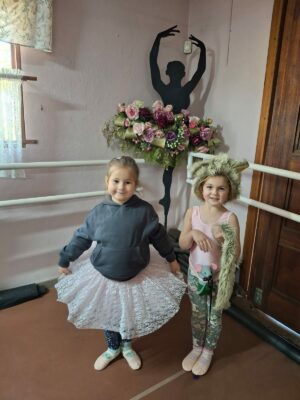 Two young girls stand in front of a ballet-themed decor. One wears a lace tutu and sweatshirt; the other a playful animal ear hat, smiling warmly.