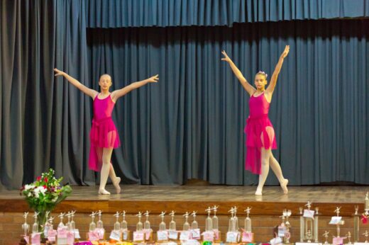 Two young dancers in pink dresses perform on stage with dark blue curtains. Trophies and flowers are lined up on the edge of the stage.