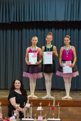 Three young ballet dancers in pink and black outfits stand on stage holding certificates, wearing medals, with a woman smiling in front; trophies displayed below.