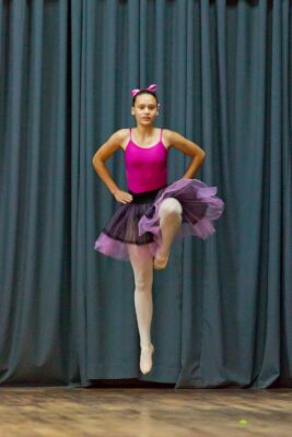 A young ballet dancer mid-jump, wearing a pink leotard with a purple tutu and matching headband, set against dark stage curtains. Focused and graceful.