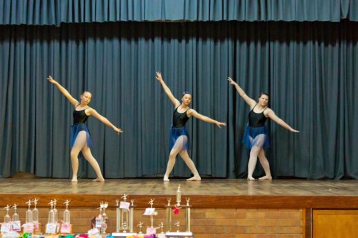 Three dancers in black leotards and blue skirts perform graceful poses on stage, with awards visible in the foreground, against a dark curtain backdrop.