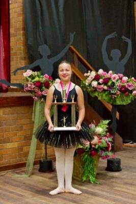 Young ballerina in a black tutu holds a large trophy, smiling in front of floral arrangements and dancer silhouettes, conveying joy and achievement.