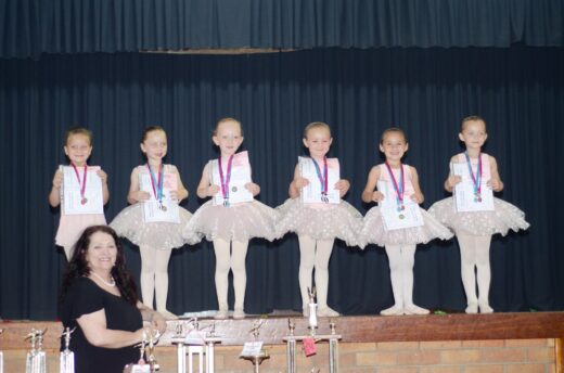 Six young girls in sparkling ballet dresses stand proudly on stage, each holding a certificate and medal. A woman smiles in front, with trophies displayed.