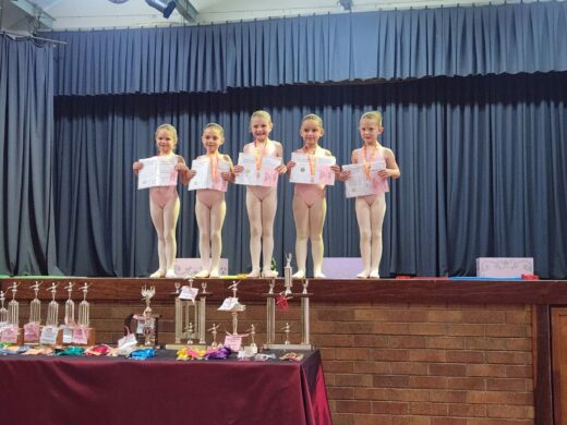 Five young ballet dancers in light pink leotards and tights stand on a stage holding certificates. Trophies and awards are displayed on a table in front of them.