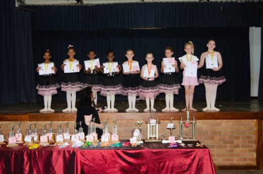 Eight young girls in ballet attire stand on stage, proudly holding certificates and medals. A table with trophies and awards is in the foreground. The scene conveys joy and accomplishment.
