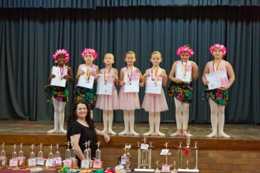 Seven young ballet dancers stand on stage, smiling and holding certificates and medals. They wear pink tutus or floral costumes. Trophies are displayed in front.