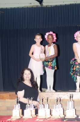 Young dancers in pink and floral costumes smile on stage holding a trophy. A woman stands in front with more trophies on a table, conveying a celebratory mood.