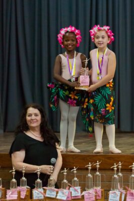 Two young ballet dancers in floral skirts and pink flower crowns hold a trophy, smiling proudly. A woman with a microphone stands in front of a table with more trophies.